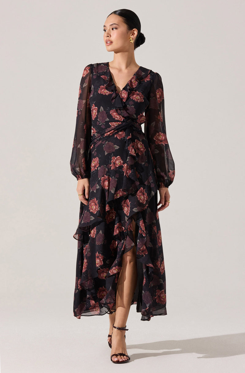 A woman is wearing a long, flowy black dress with a floral pattern featuring pink and purple flowers. The dress has long, sheer sleeves and a ruffled, layered skirt with a slit in the front. The neckline is decorated with a ruffle trim.