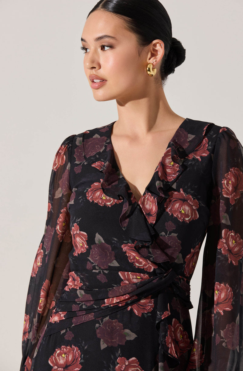 A woman wearing a black dress with a red and pink floral pattern. The dress features a ruffled neckline, long sheer sleeves, and a waist tie that creates a gathered effect at the waist.