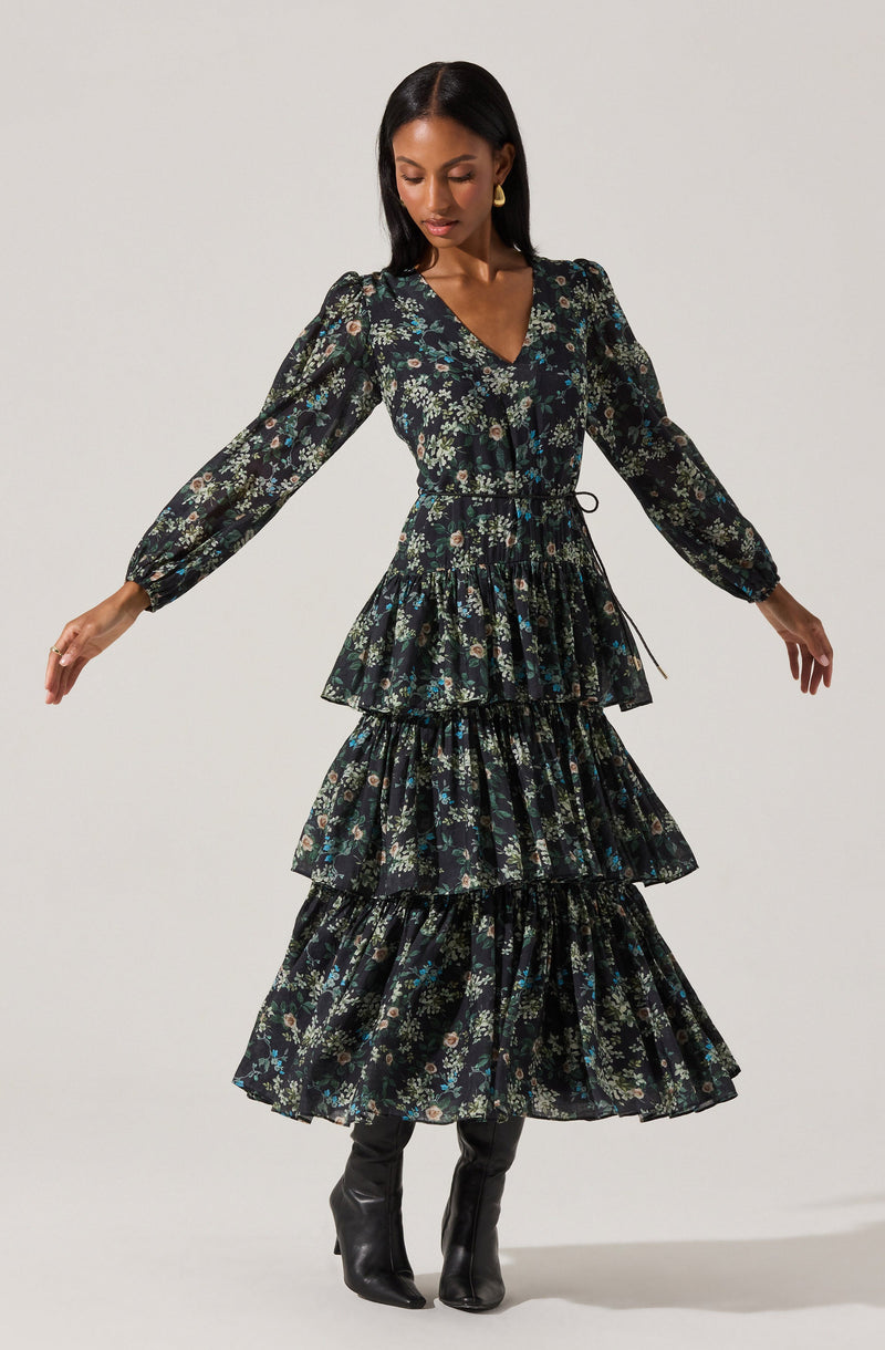 A woman is wearing a long, dark floral dress with a V-neckline and long, puffed sleeves. The dress features a fitted waist and three tiered, ruffled skirt sections. She pairs the dress with black knee-high boots and gold earrings.