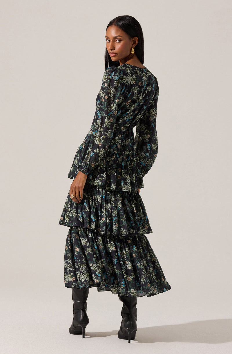 A woman is wearing a long, black dress with a floral pattern featuring small flowers in shades of white, blue, and green. The dress has long sleeves with elastic cuffs, a cinched waist, and three-tiered ruffled skirts. She is also wearing black knee-high boots with pointed toes and stiletto heels, and she has gold hoop earrings.