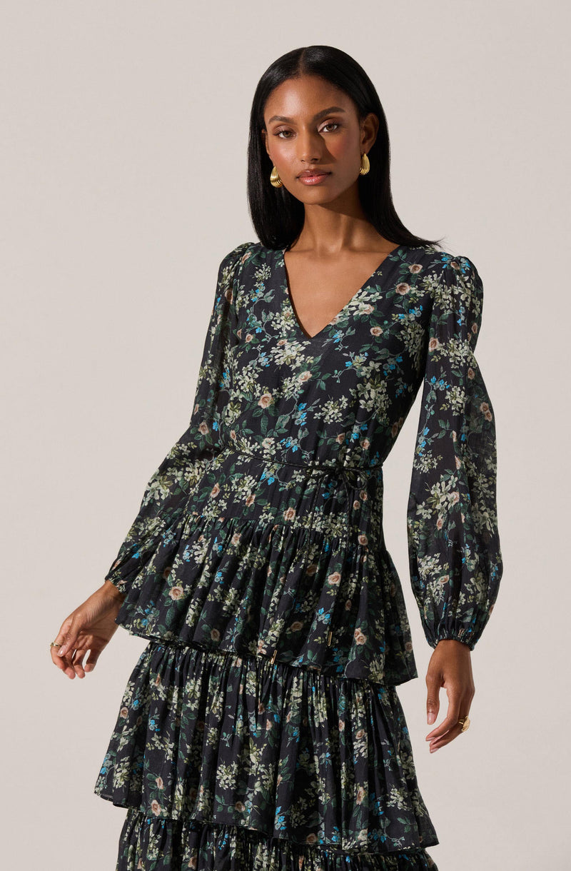 A woman is wearing a long black dress with a floral pattern. The dress features a V-neckline, long puffed sleeves with gathered cuffs, and a tiered skirt with multiple ruffled layers. The floral pattern includes various small flowers and leaves in shades of green, cream, pink, and blue.