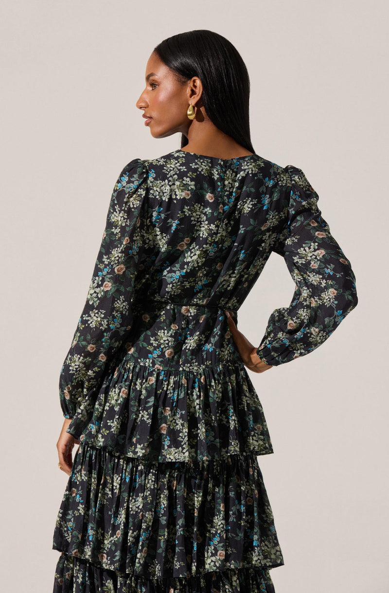 A woman is wearing a long-sleeved dress with a dark background and a floral pattern featuring small flowers in shades of green, pink, and blue. The dress has a fitted waist, puffed shoulders, and a tiered, ruffled skirt. She is accessorizing with large, gold hoop earrings and has her hair styled straight and sleek.