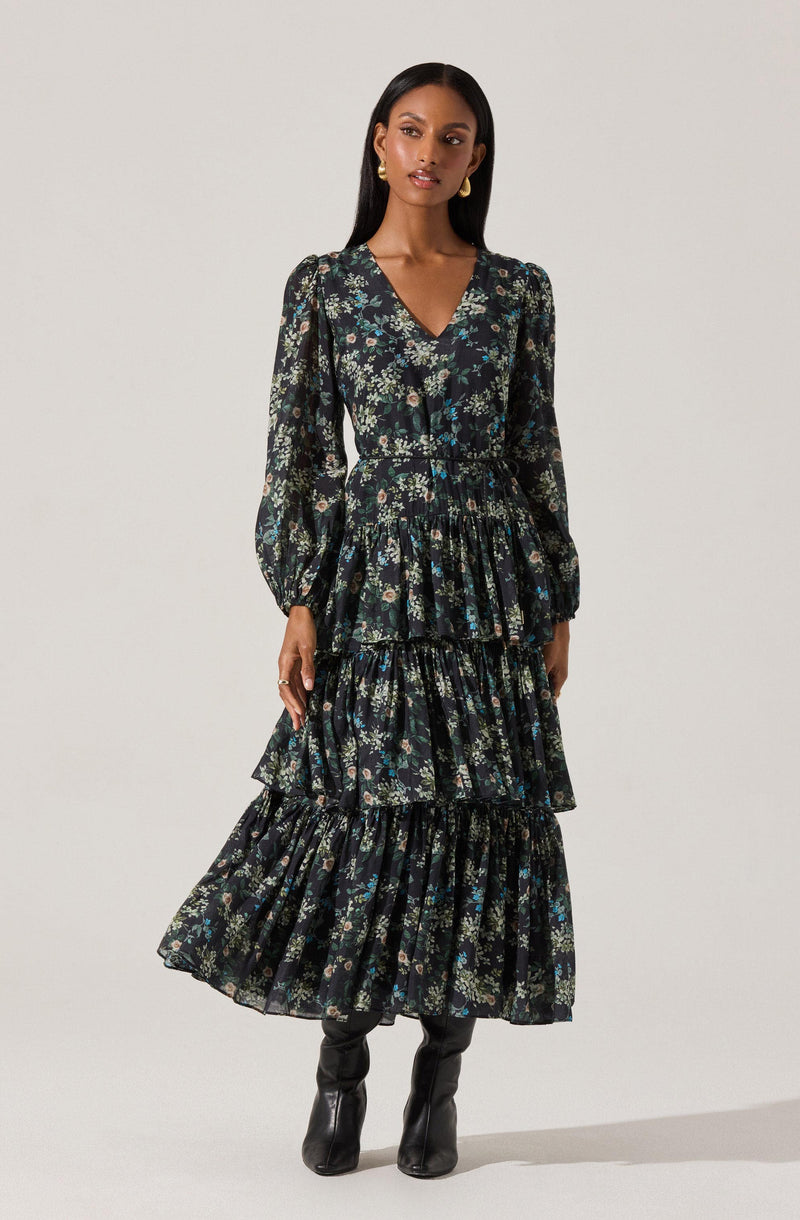 A woman is wearing a long, black floral dress with a V-neckline. The dress features a tiered skirt with multiple ruffles and long, slightly puffed sleeves. The floral pattern includes small flowers in shades of blue, green, and beige. She completes her outfit with black knee-high boots and gold earrings.