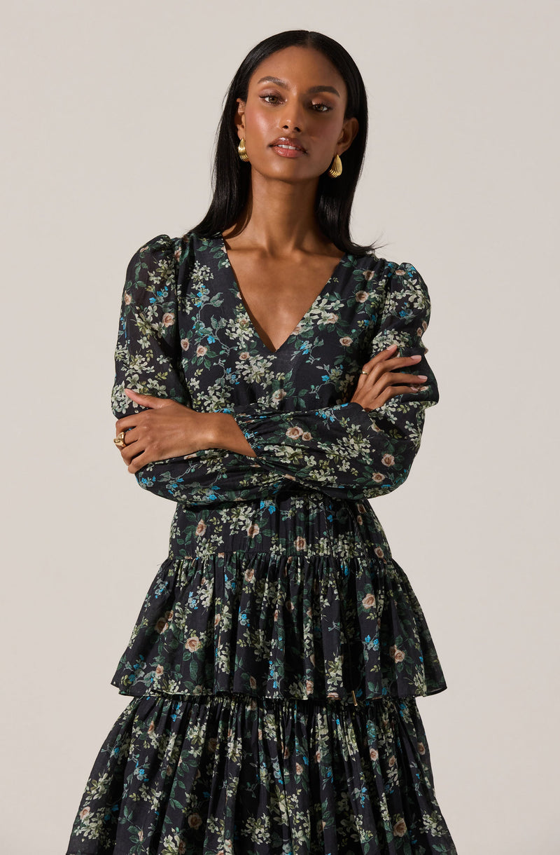 A woman is wearing a long, flowing dress with a black background and a floral pattern featuring small flowers in shades of blue, beige, and green. The dress has a V-neckline, long puffed sleeves, and a tiered skirt with ruffled layers. She accessorizes with gold hoop earrings and a gold ring.
