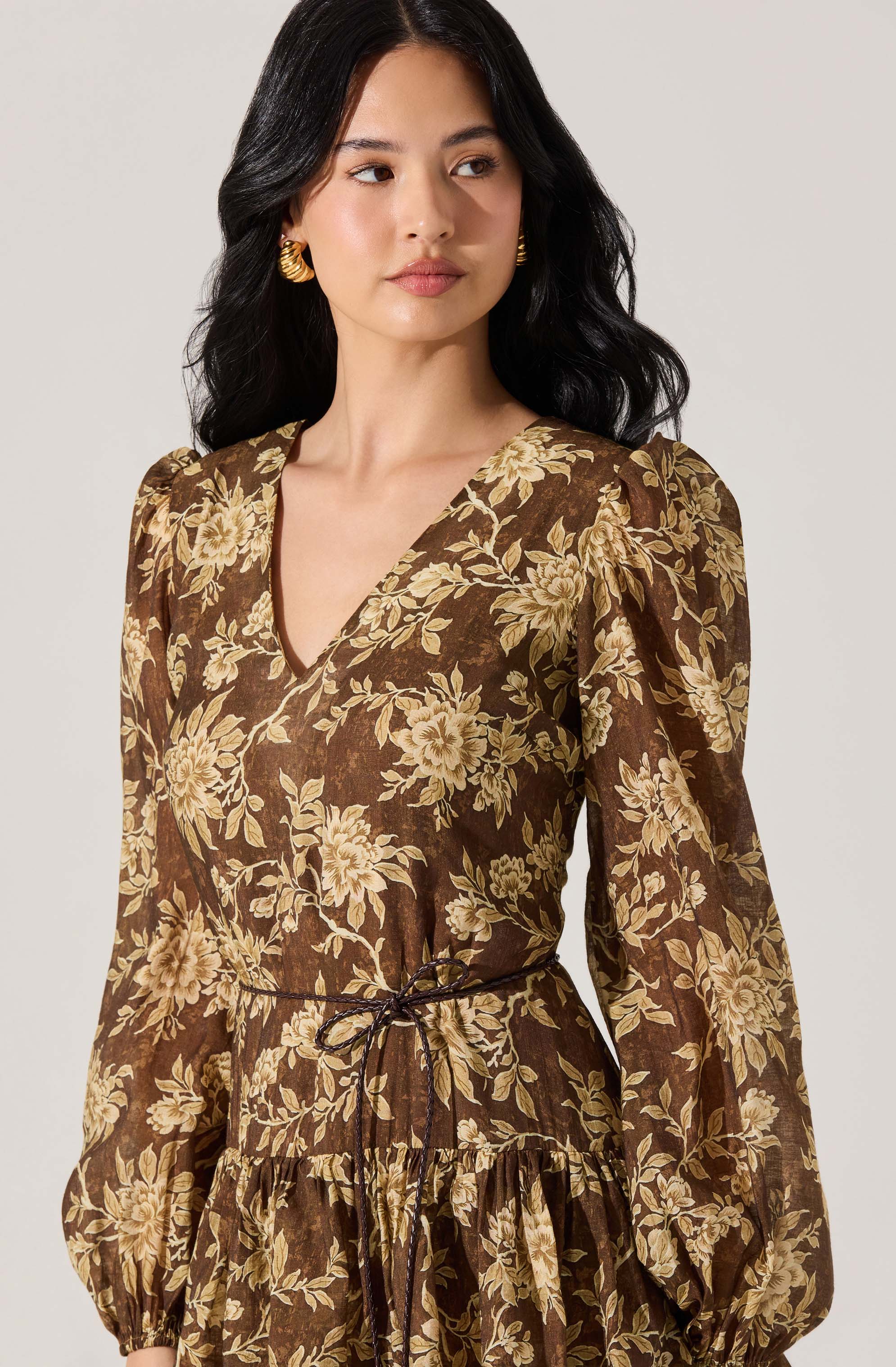 A woman is wearing a long-sleeved dress with a V-neckline and a floral print in shades of beige and cream on a brown background. The dress features puffed shoulders, a gathered waist with a thin tie, and a ruffled skirt.