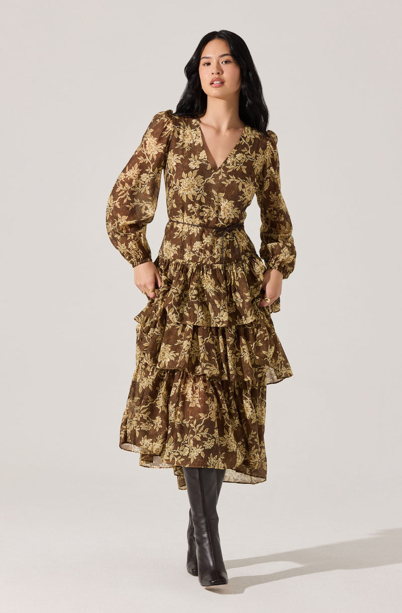 A woman is wearing a long, brown dress with a floral pattern in shades of beige. The dress has a V-neckline, long puffed sleeves, a fitted waist, and multiple tiered ruffles that extend down to her ankles. She is also wearing black knee-high boots.