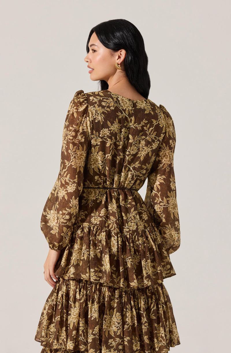 A woman is wearing a long dress with a brown background and an intricate gold floral pattern. The dress has long, puffed sleeves with elastic cuffs, a fitted waist, and a tiered skirt with ruffles.