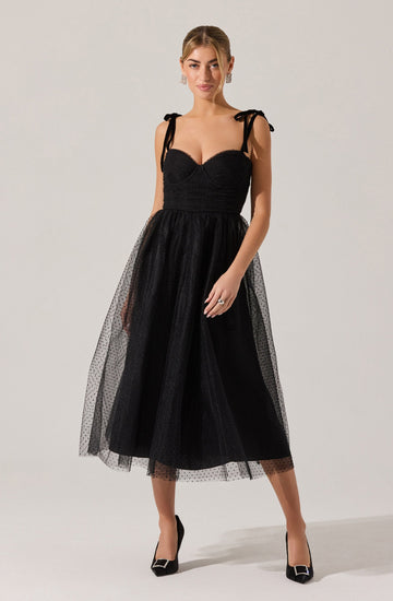 A woman is wearing a black dress with a fitted, ruched bodice featuring sweetheart neckline and shoulder straps tied into bows. The dress has a high waistline and a full, sheer tulle overlay skirt that falls to mid-calf length. She is also wearing black high heels with a decorative buckle detail on the toe.