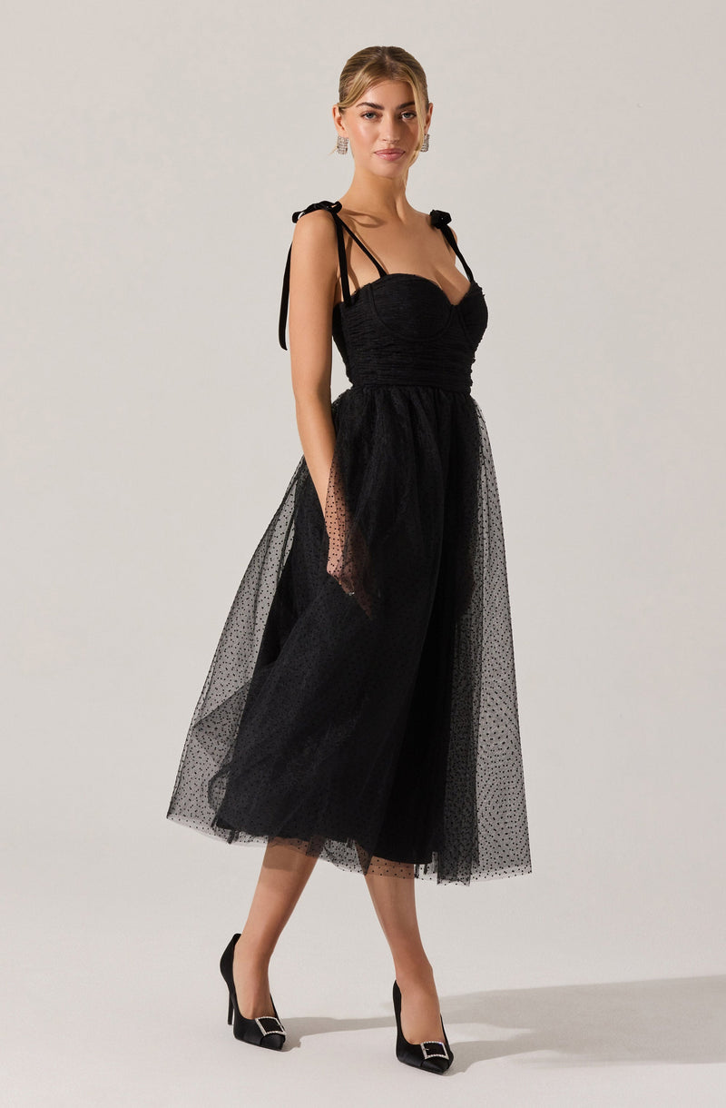 A woman is wearing a black dress with a fitted, ruched bodice and thin shoulder straps tied into bows. The skirt is made of a sheer, polka-dotted tulle fabric, creating a layered, voluminous look that reaches mid-calf. She pairs the dress with black high-heeled shoes featuring pointed toes and metallic embellishments on the front.