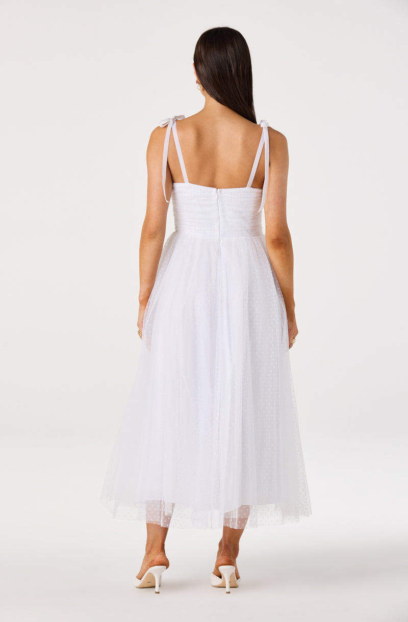 Woman in a white, sleeveless dress with shoulder tie straps and tulle skirt, wearing white heels.