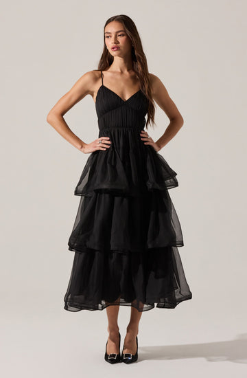 A woman is wearing a black dress with a fitted bodice and thin spaghetti straps. The dress features multiple tiers of voluminous, sheer fabric that create a layered and ruffled effect from the waist down to the hem. She pairs the dress with black high heels that have a pointed toe.