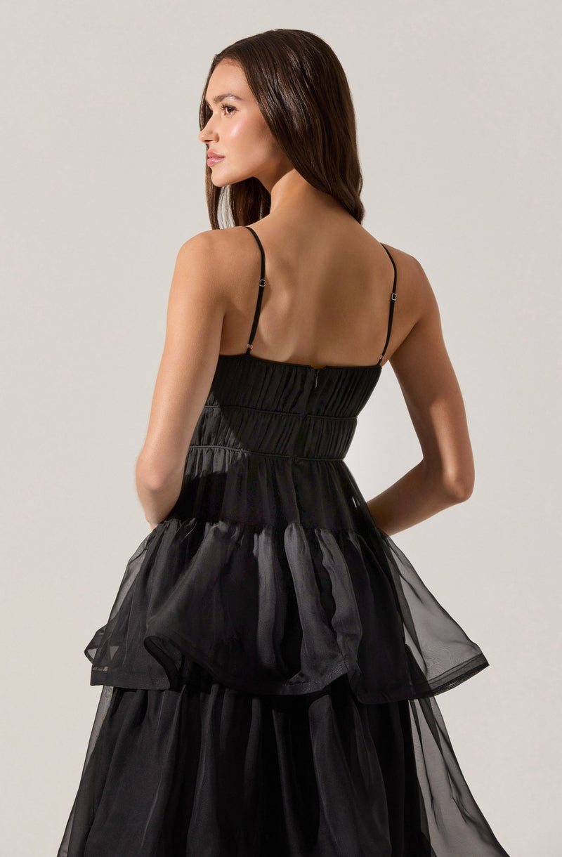 A woman is wearing a sleeveless black dress with thin adjustable spaghetti straps. The dress has a fitted, ruched bodice and a layered, voluminous skirt made of sheer, flowing fabric.