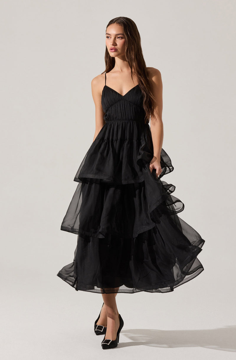 A woman is wearing a black dress with thin spaghetti straps, a fitted bodice with gathered fabric detail, and a voluminous, tiered tulle skirt that reaches mid-calf. She is also wearing black high heels with a rounded square buckle on the toe.