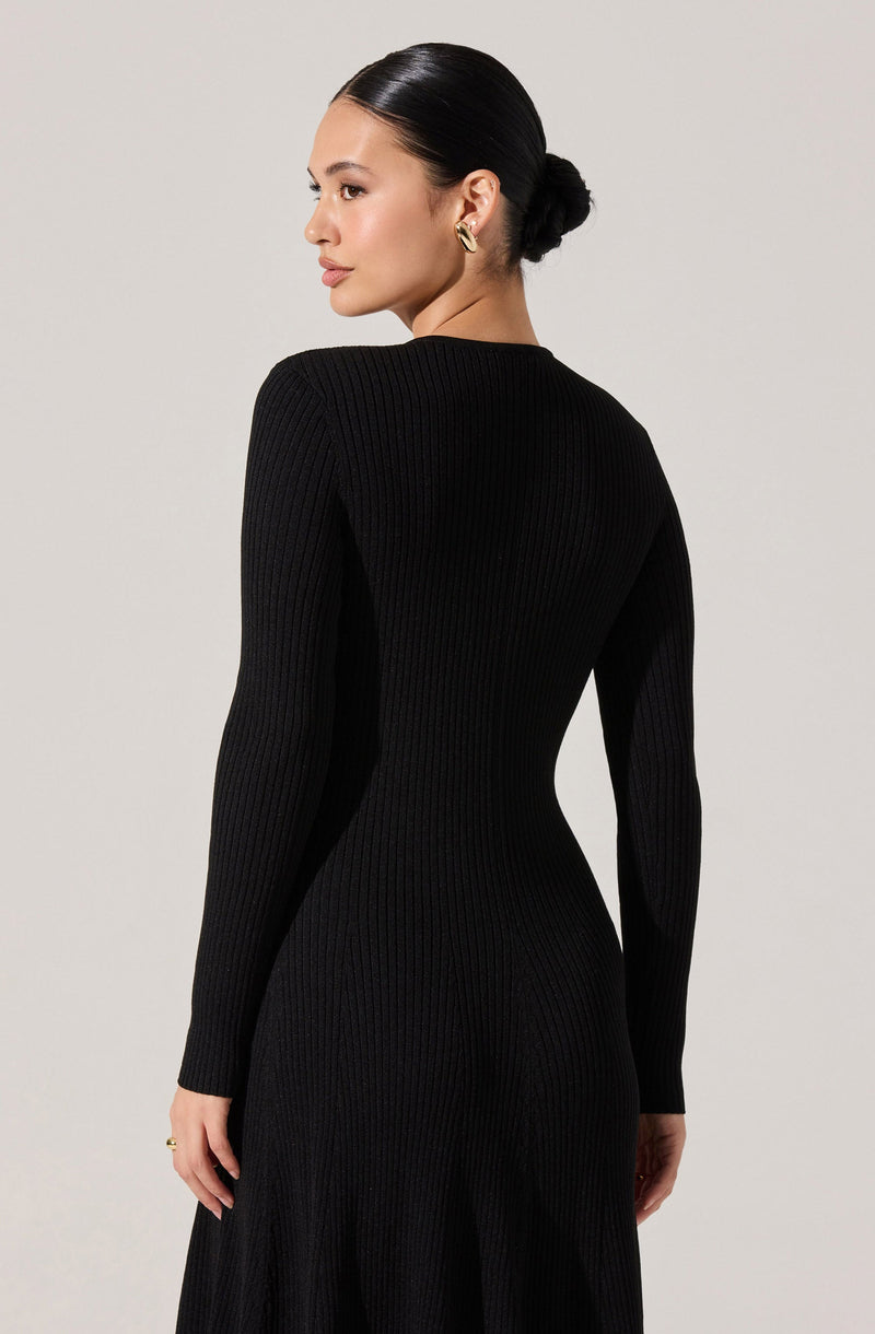 A woman is wearing a form-fitting, long-sleeved, black ribbed knit dress with a round neckline. The dress emphasizes her waist and has a subtle flare at the skirt's hem.
