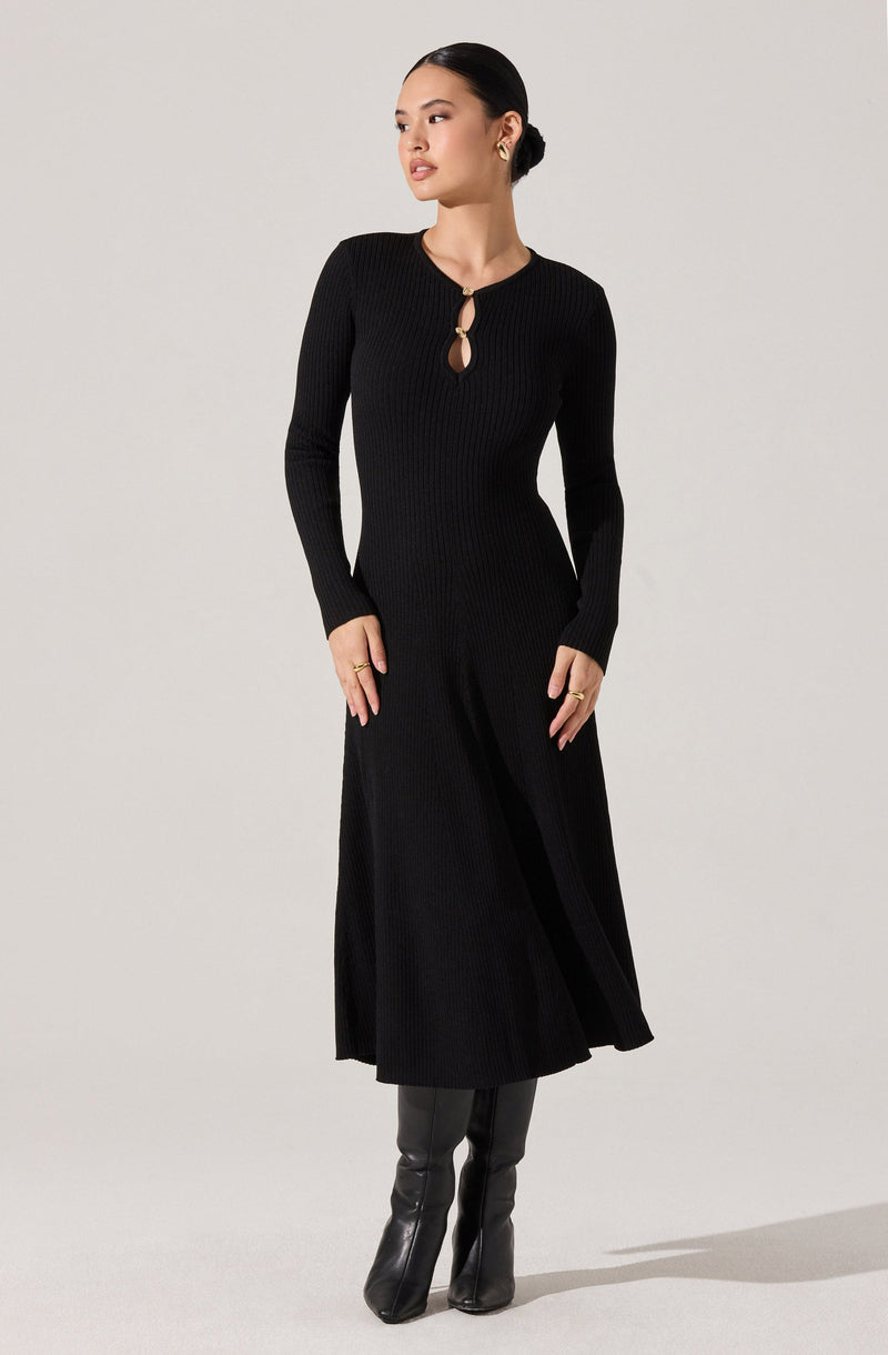 A woman is wearing a long, black, ribbed knit dress with a round neckline and a small keyhole opening secured by a circular knot detail. The dress has long sleeves and a fitted waist, flowing into an A-line skirt that reaches her ankles. She is also wearing black knee-high boots.