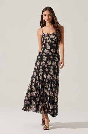 A woman is wearing a long, flowy black dress with a floral pattern featuring white and pink flowers. The dress has spaghetti straps and a fitted bodice that flares out at the bottom.