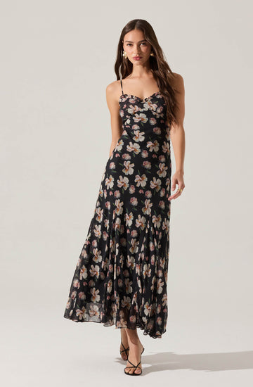 A woman is wearing a long, flowy black dress with a floral pattern featuring white and pink flowers. The dress has spaghetti straps and a fitted bodice that flares out at the bottom.