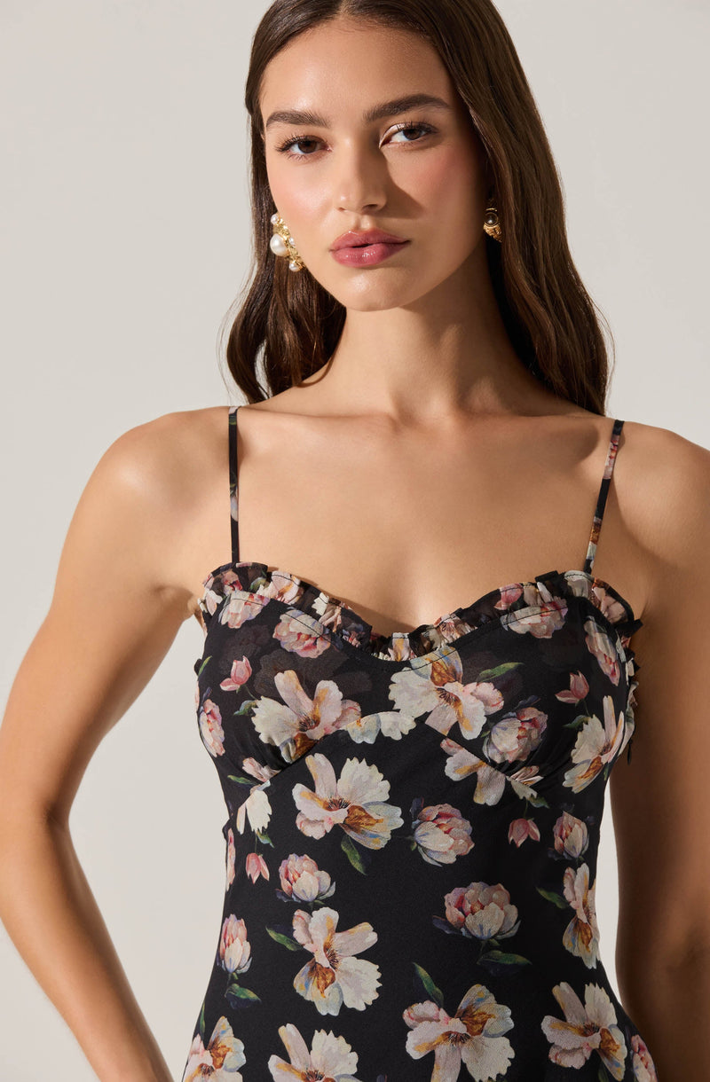 A woman is wearing a black sleeveless dress with thin spaghetti straps. The dress features a floral pattern of large, light-colored flowers with pink and beige petals and green leaves. The top of the dress has a ruffled detail along the neckline. She is accessorized with large, statement earrings that have a pearl and gold design.