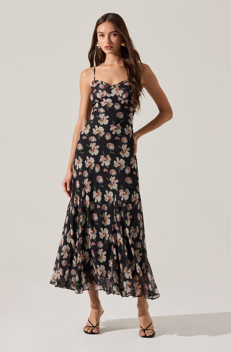 A woman is wearing a sleeveless, black maxi dress with thin spaghetti straps. The dress features a floral pattern with large white and pink flowers scattered across the fabric. The hem of the dress is ruffled, adding a flowing, feminine touch.
