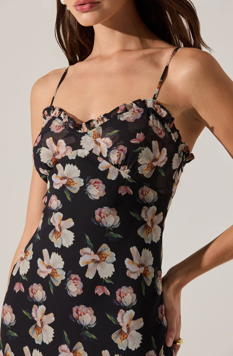 A woman is wearing a black dress with a floral pattern featuring large, light-colored flowers with pink, white, and orange petals, and green leaves. The dress has thin, spaghetti straps and a ruffled trim along the neckline.