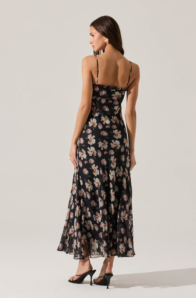 A woman is wearing a black, sleeveless maxi dress with thin spaghetti straps. The dress features a floral pattern with large, light-colored flowers scattered throughout. She is also wearing high-heeled sandals.