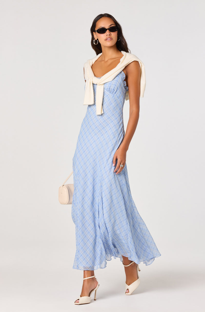 Woman wearing a light blue plaid maxi dress with ruffles, cream cardigan tied around shoulders, black sunglasses, and white high heels.