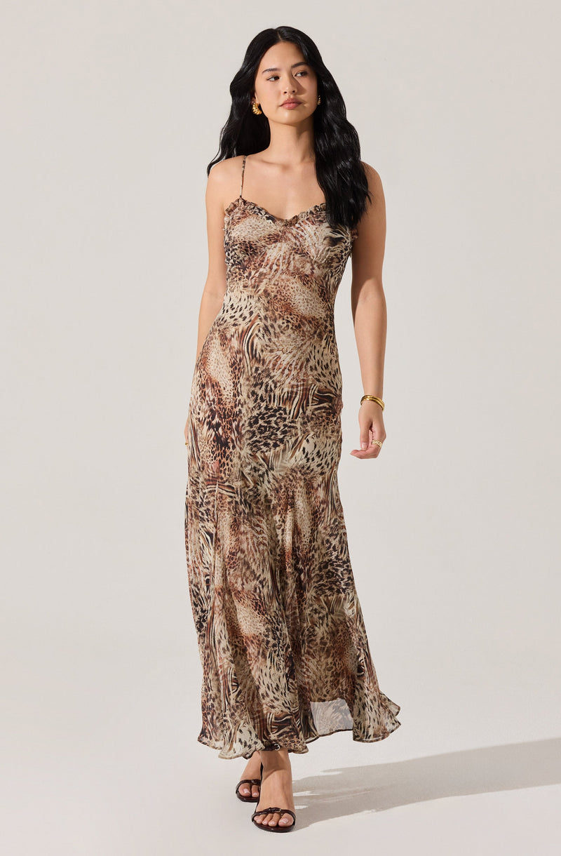 A woman wearing a long, flowing dress with a leopard print pattern. The dress features thin spaghetti straps and a slightly ruffled neckline. She is also wearing gold jewelry, including earrings and a bracelet. The overall look is elegant and stylish.