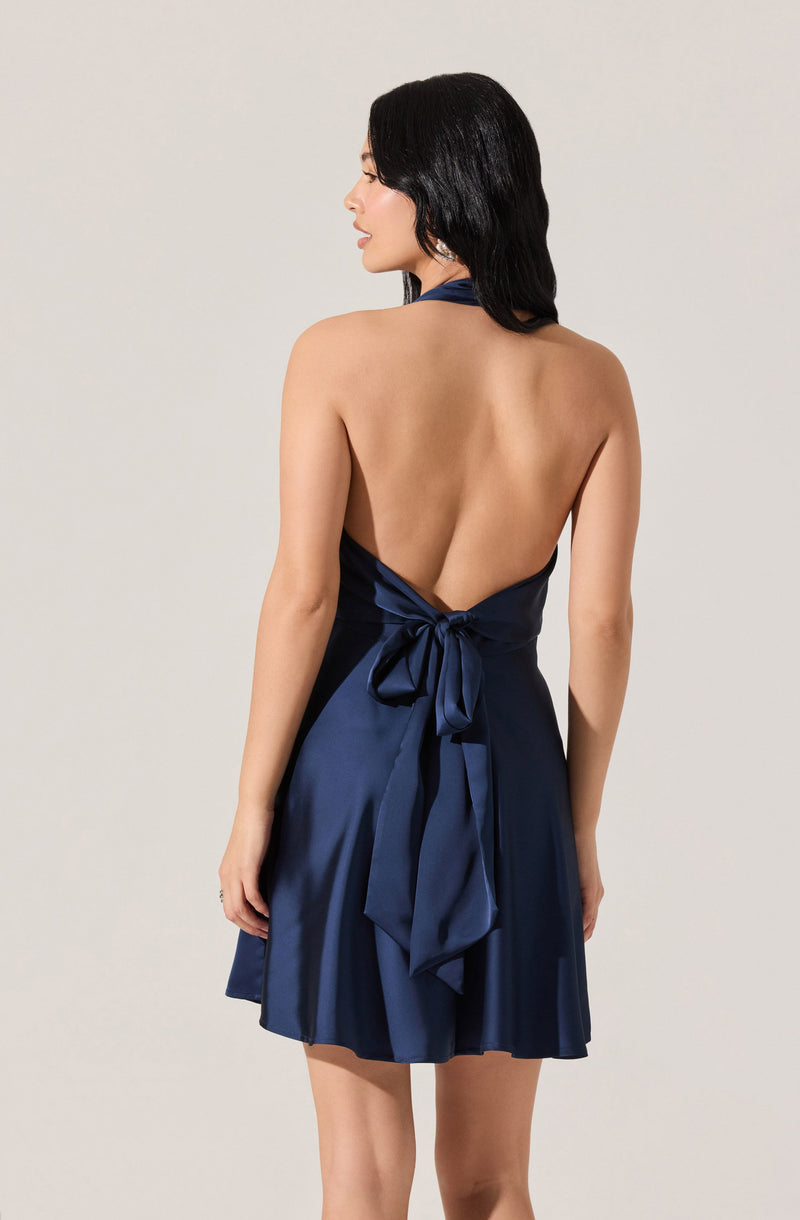 A woman is wearing a sleeveless, dark blue satin dress with a halter neckline that ties into a large bow at the back. The dress has an open back and a short, flowy skirt.