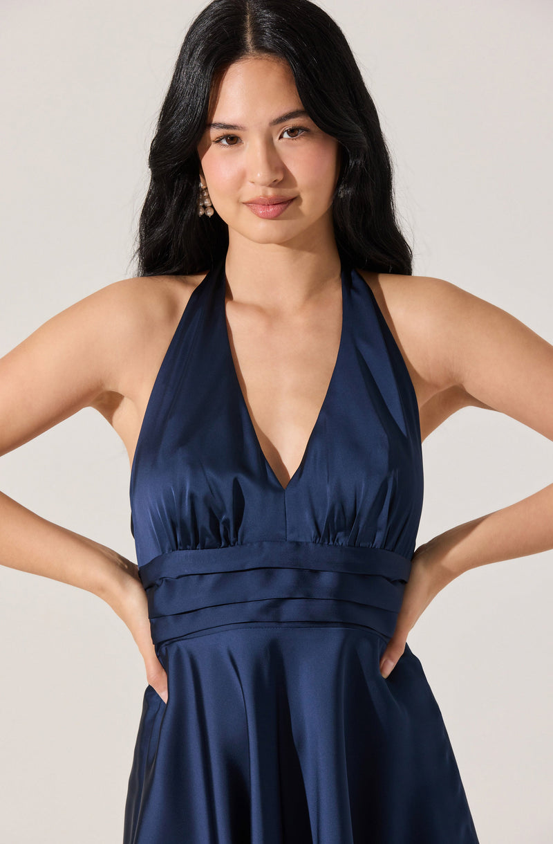 A woman is wearing a deep blue satin halter dress with a V-neckline. The dress features gathered fabric under the bust and horizontal pleats at the waist, creating a fitted bodice. The satin material has a smooth, shiny finish, giving the dress an elegant appearance.