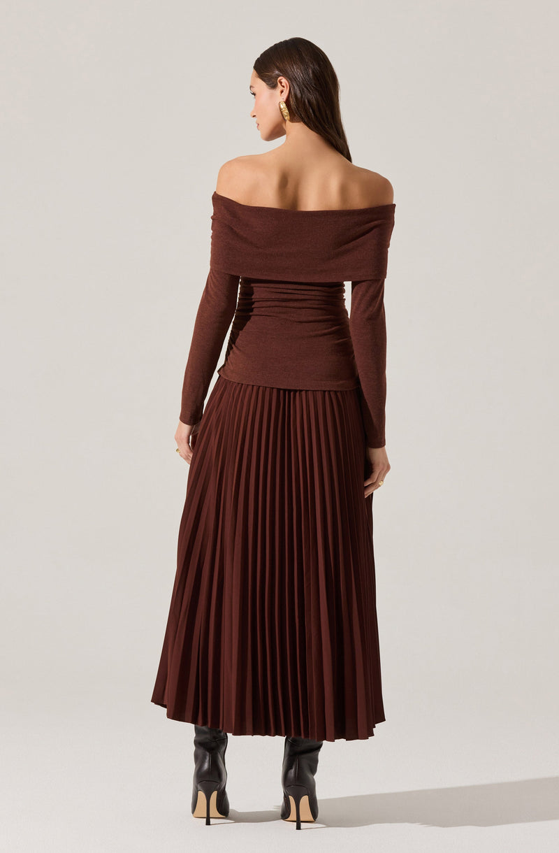 A woman is wearing an off-the-shoulder, long-sleeve, deep brown fitted top with a wide neckline that drapes across her shoulders. She pairs it with a high-waisted, pleated, dark brown midi skirt that reaches below her calves. She completes her outfit with tall black heeled boots.