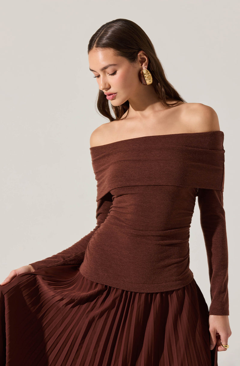A woman is wearing an off-the-shoulder, long-sleeved top in a rich brown color with a wide, folded neckline that sits across her shoulders. She pairs it with a matching pleated skirt made of a shiny fabric. She accessorizes with large, statement gold earrings.