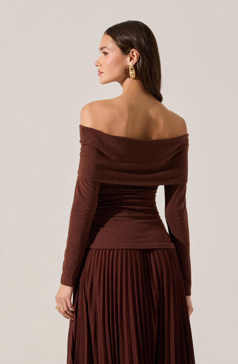 A woman is wearing an off-the-shoulder, long-sleeved, fitted brown top with a wide neckline that drapes over her shoulders. She has on a large textured gold hoop earring in each ear. Her hair is straight and dark, styled behind her ears.