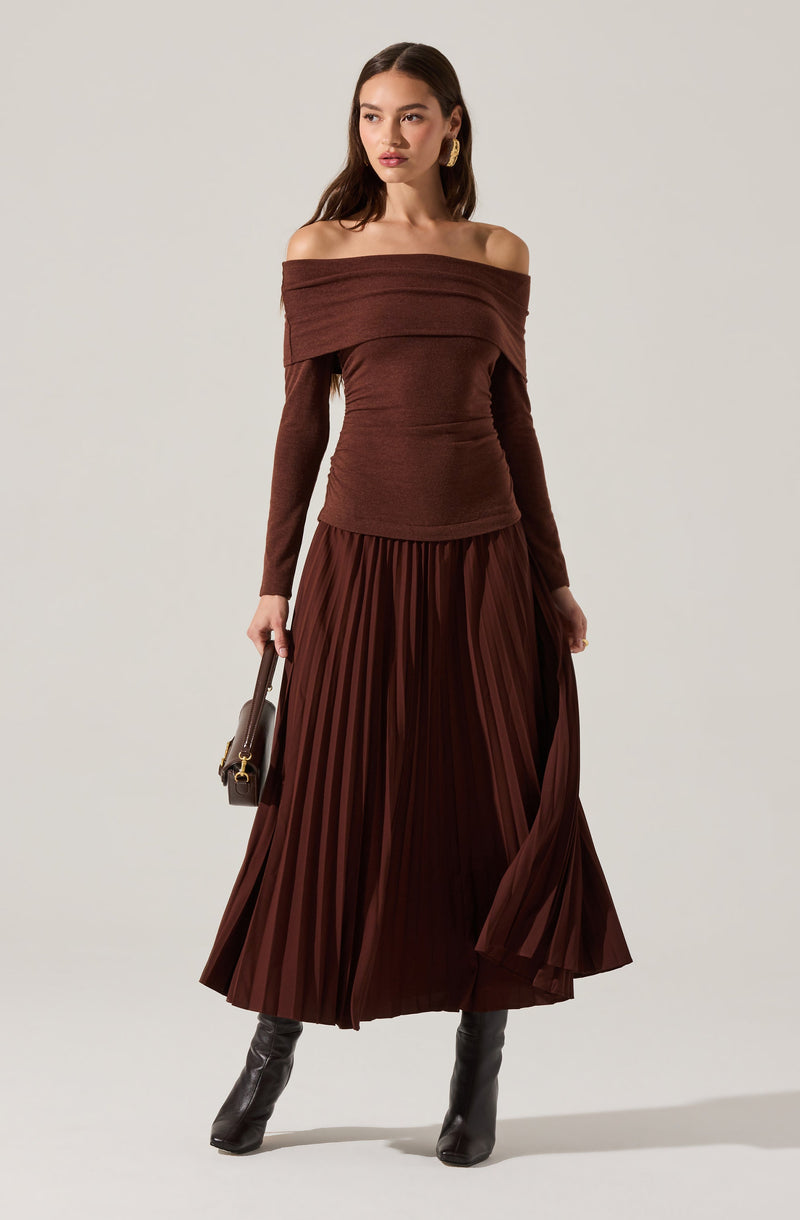 A woman is wearing an off-the-shoulder, long-sleeved brown top with a wide neckline. She pairs it with a matching high-waisted, pleated, floor-length skirt in a rich brown color. She accessorizes with large gold hoop earrings, black knee-high boots, and carries a small dark handbag with a short handle.
