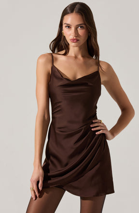 A woman wearing a sleeveless, dark brown satin dress with thin spaghetti straps. The dress features a cowl neckline and a draped, slightly asymmetrical hem.