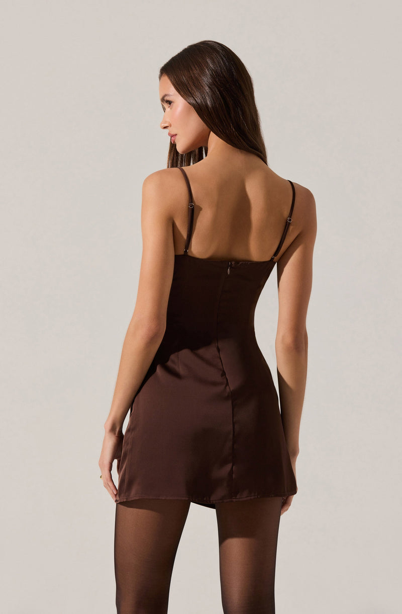 A woman is wearing a fitted satin mini dress in a dark chocolate color with spaghetti straps and a back zipper. She is also wearing sheer black tights.
