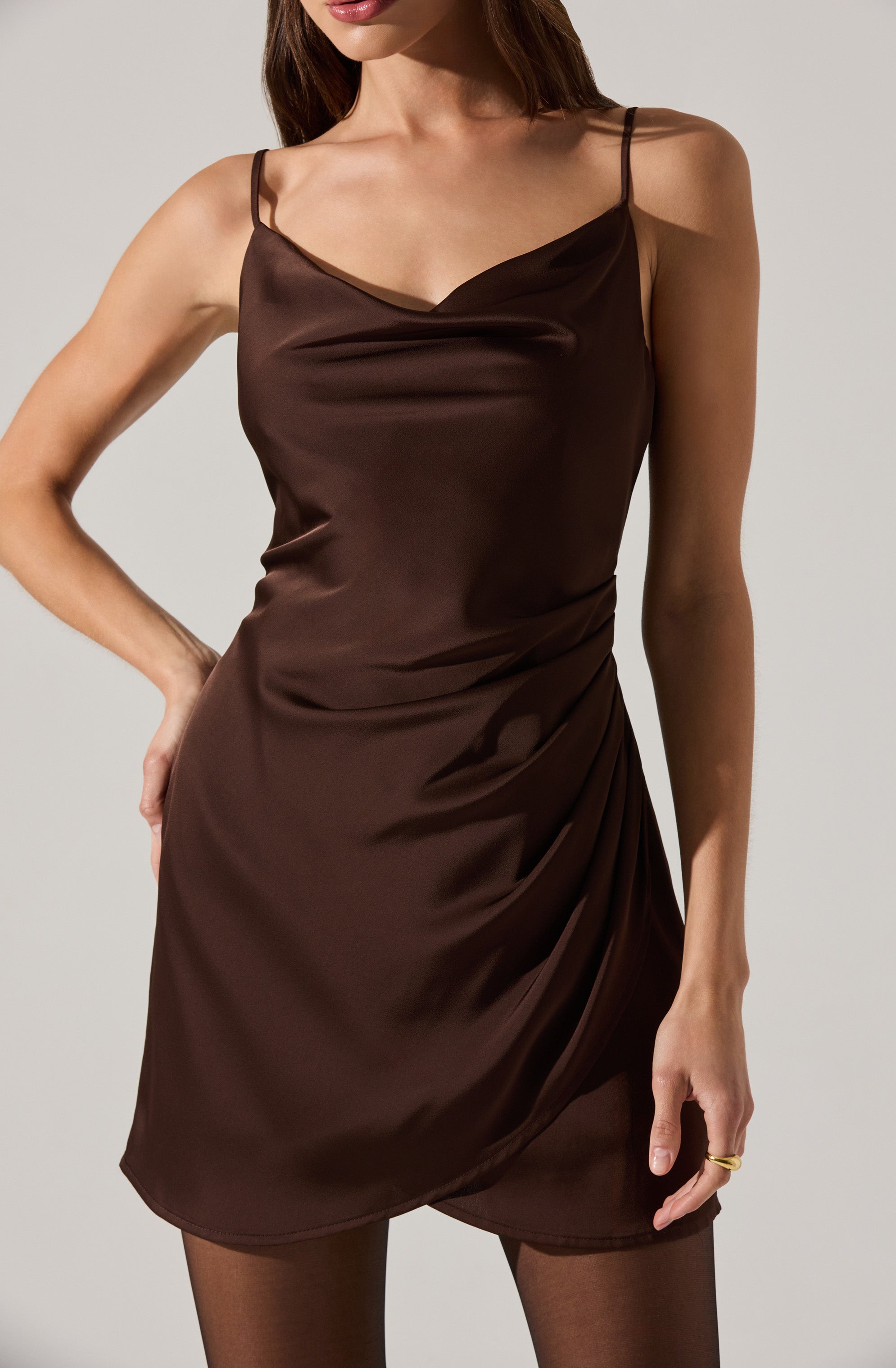 A woman is wearing a sleeveless, dark brown satin dress with thin spaghetti straps. The dress has a cowl neckline and features draped fabric details on the side, creating a gathered effect. She is also wearing sheer black tights.