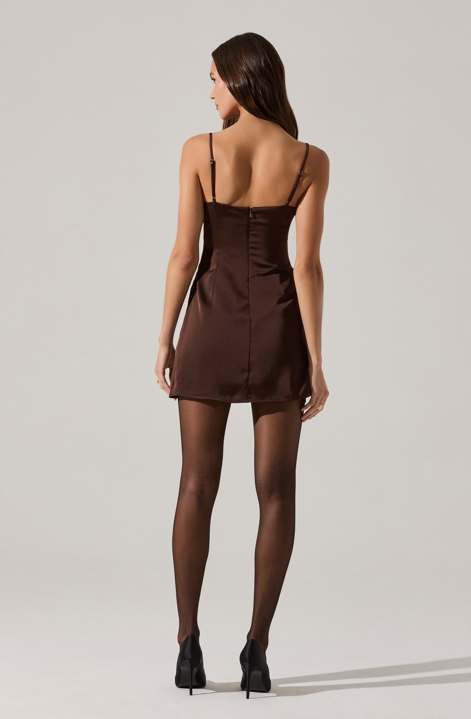 A woman is wearing a short, dark brown satin dress with thin spaghetti straps and a square neckline at the front. She pairs the dress with sheer black tights and black high heels.