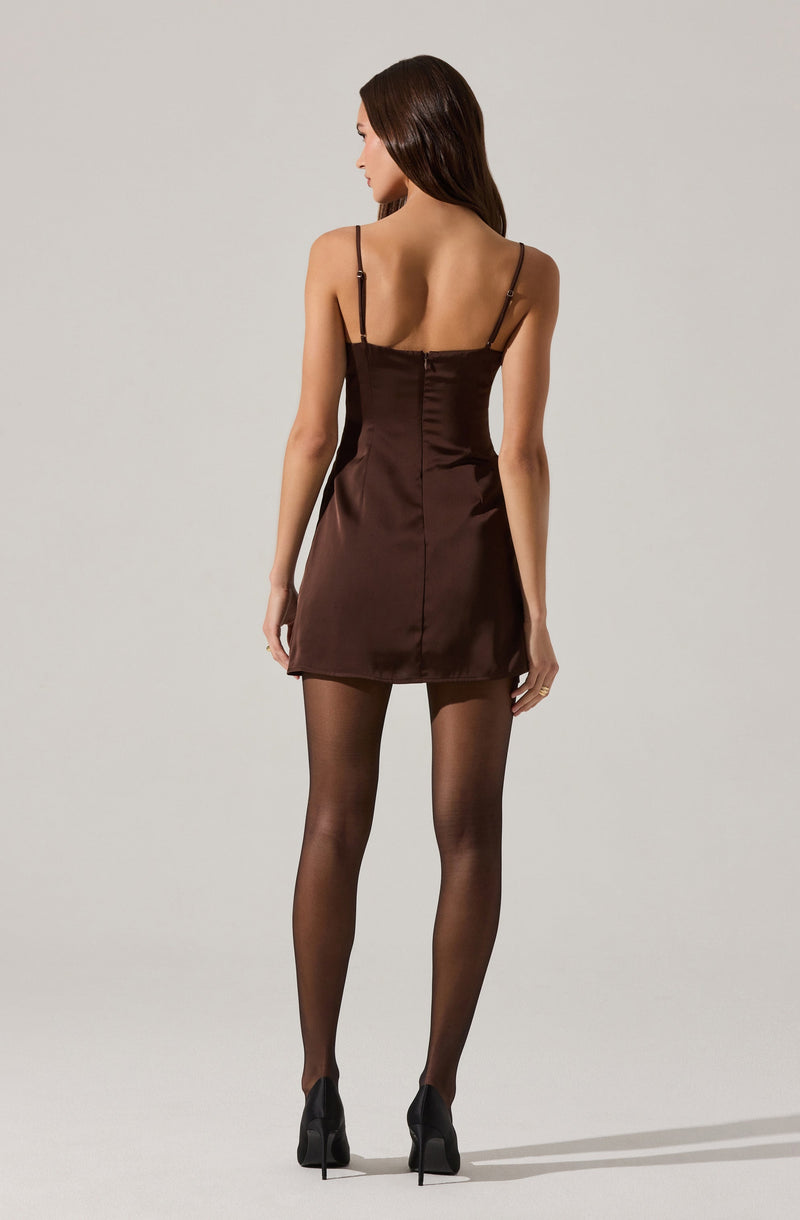A woman is wearing a short, dark brown satin dress with thin spaghetti straps and a square neckline at the front. She pairs the dress with sheer black tights and black high heels.