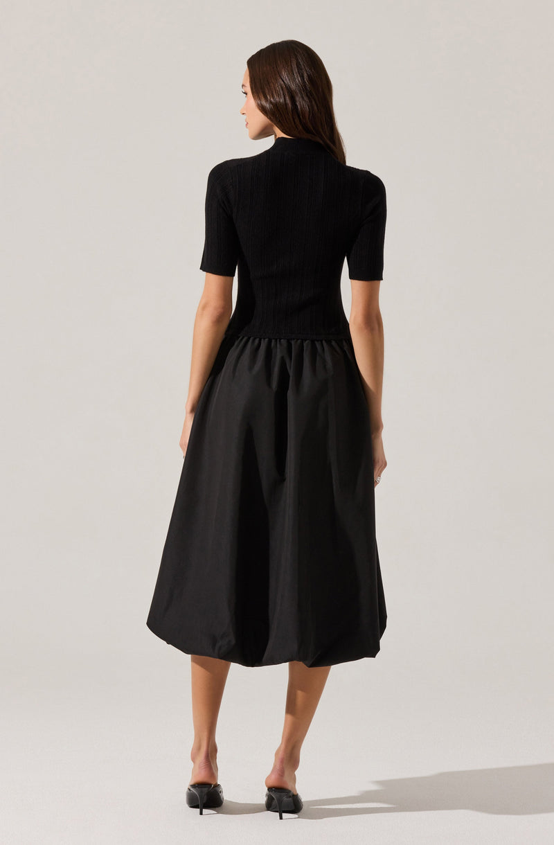 A woman is wearing a fitted, short-sleeved black top made of ribbed fabric, paired with a voluminous, knee-length black skirt that has a gathered waist. She is also wearing black high-heeled shoes.