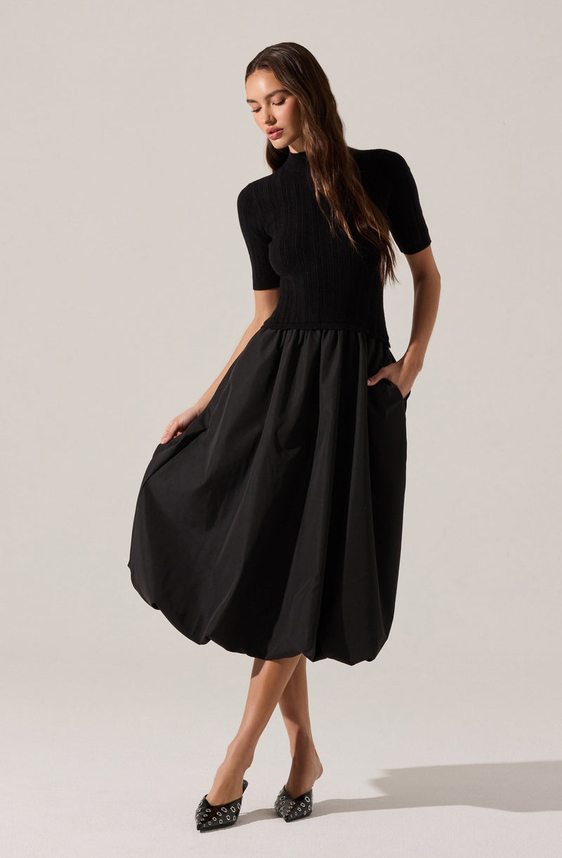 The woman is wearing a black short-sleeved knit top with a high neckline, fitted to her torso. She is also wearing a voluminous black midi-length skirt with a gathered waist and pockets, made of a shiny, structured fabric that creates a bubble-like shape. Her shoes are black pointed-toe heels decorated with metallic embellishments.