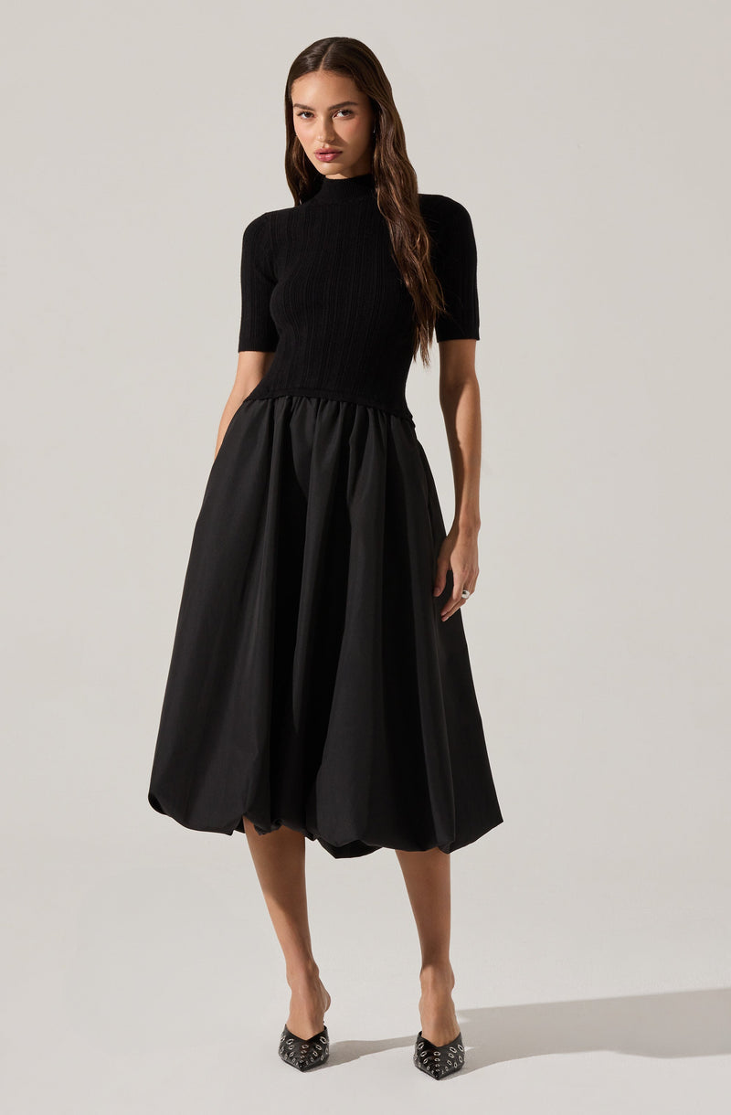 A woman is wearing a fitted, short-sleeved, black ribbed turtleneck top paired with a voluminous, midi-length black skirt that has a gathered, bubble design. She completes the look with pointed high heels that have a black and white pattern.