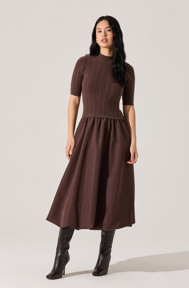 A woman is wearing a fitted, short-sleeved, ribbed knit top in a dark brown color with a high neckline. She pairs it with a voluminous, ankle-length skirt in a matching dark brown satin fabric, creating a monochromatic outfit. She completes her look with a pair of tall, black heeled boots.