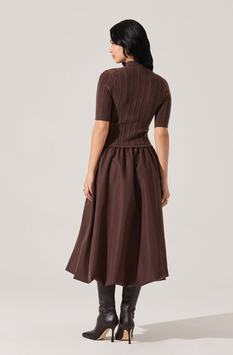A woman is wearing a fitted, short-sleeved, ribbed knit top in a dark brown color. She pairs it with a high-waisted, full-length, dark brown silk or satin skirt that has a gathered waist. She completes the outfit with tall, black leather stiletto boots.