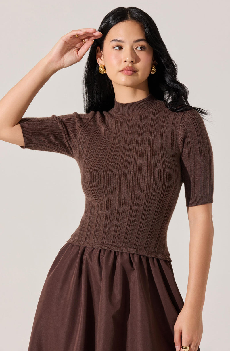 A woman is wearing a fitted, ribbed, short-sleeve knit top in a rich brown color, with a high neckline. She pairs the top with a full, gathered skirt in a matching brown shade, made of a shiny, smooth fabric. She is accessorized with large, gold, textured earrings.