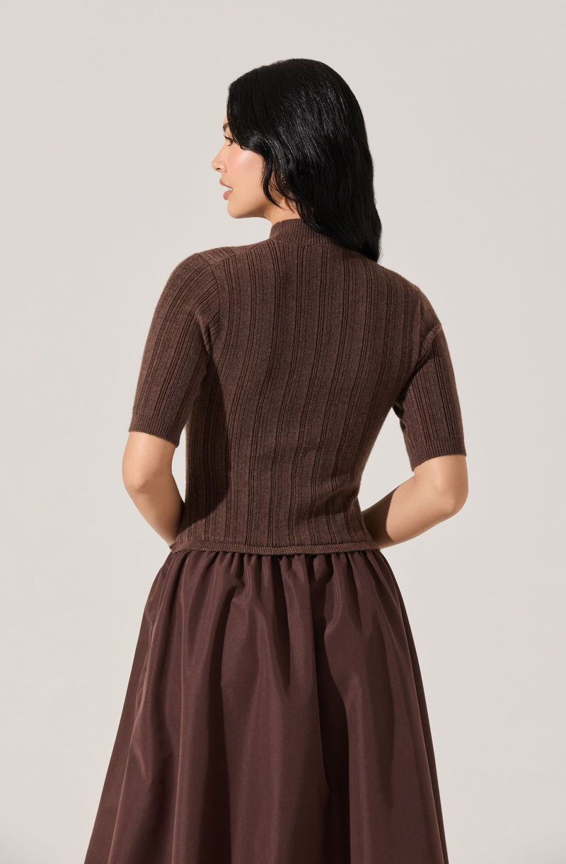 A woman is wearing a fitted, short-sleeved, ribbed brown sweater that reaches her hips. She pairs it with a full, high-waisted skirt made of shiny fabric in a similar brown shade. Her hair is long, black, and wavy.