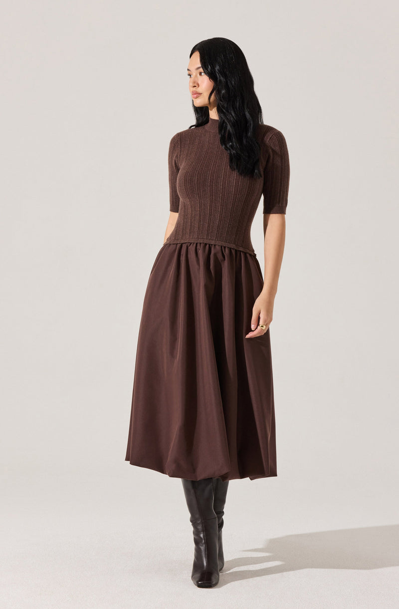 A woman is wearing a fitted, short-sleeved, ribbed brown top with a high neckline. She pairs it with a high-waisted, full-length, dark brown skirt that has a gathered, slightly puffed design. She is also wearing black knee-high boots.