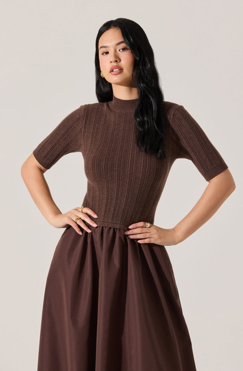 A woman is wearing a fitted, short-sleeved, ribbed brown sweater with a high neckline. She pairs it with a full, high-waisted, dark brown satin or silk skirt that has a gathered, slightly pleated design. She has long, wavy black hair and is accessorized with gold earrings and rings.