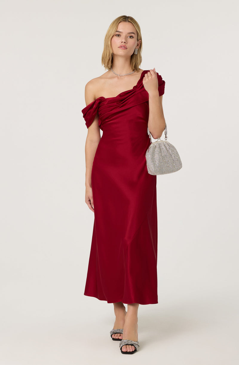 A woman is wearing a long, elegant, off-the-shoulder velvet gown in a deep red color. The dress features a gathered bodice with draped fabric on the shoulder. She accessorizes with a sparkling choker necklace, dangling earrings, and high-heeled sandals with embellishments. She is holding a small, silver, rhinestone-encrusted handbag.
