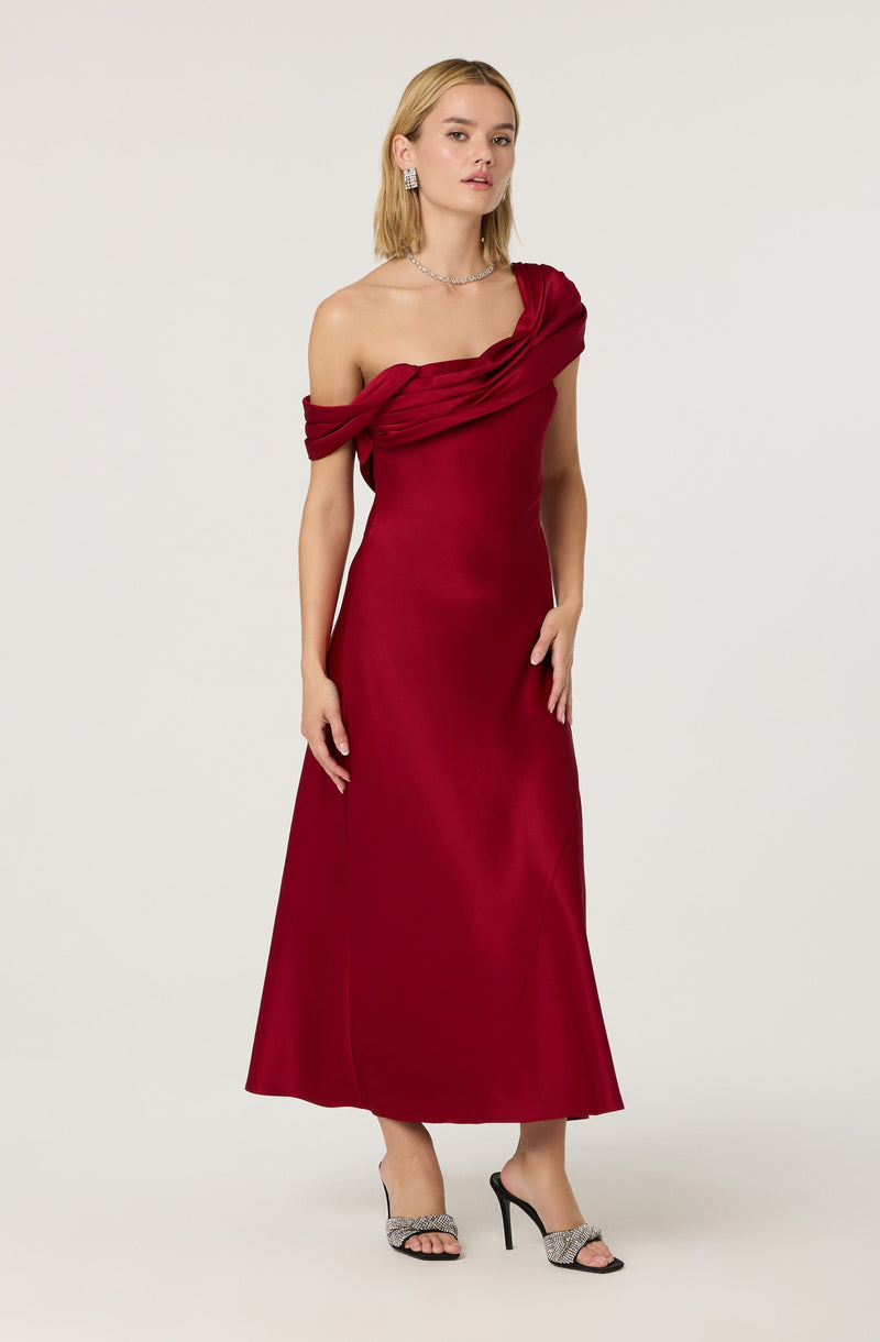 A woman is wearing a floor-length, off-the-shoulder red gown with draped fabric details around the neckline and shoulder. She is accessorized with earrings and a necklace, and is wearing high-heeled sandals with a black and white pattern.
