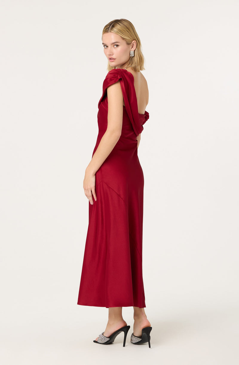 A woman is wearing a long, elegant red gown with a strapless design and a prominent ruffle detail on one shoulder. She has ankle-high, open-toe black heels with embellishments. She also has statement earrings and styled blonde hair.